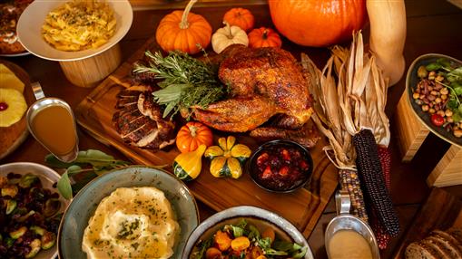 Photo shows a table filled with a Thanksgiving meal including turkey, ham, Brussels sprouts, mashed potatoes, and pumpkins.